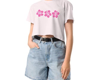 Könnte beinhalten: Ein weißes, bauchfreies T-Shirt mit einer Reihe von drei pinkfarbenen Hibiskusblüten, die auf die Vorderseite gedruckt sind. Das Shirt wird mit hellblauen Jeans-Shorts und einem schwarzen Gürtel kombiniert.