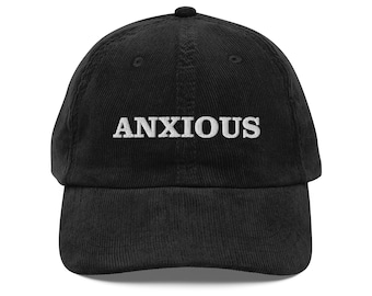 Könnte beinhalten: Eine schwarze Baseballkappe aus Kord mit dem Wort ANXIOUS, das in weißen Großbuchstaben auf der Vorderseite eingestickt ist. Die Kappe hat eine strukturierte Krone, einen gebogenen Schirm und einen Knopf oben aus strukturiertem Stoff.