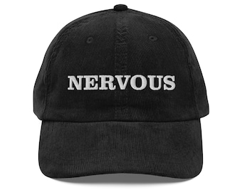 Könnte beinhalten: Eine schwarze Baseballkappe aus Cord mit dem Wort NERVOUS, das in weißen Großbuchstaben auf die Vorderseite gestickt ist. Die Kappe hat eine strukturierte Krone, einen gebogenen Schirm und einen Knopf oben, mit einer deutlich gerippten Textur.