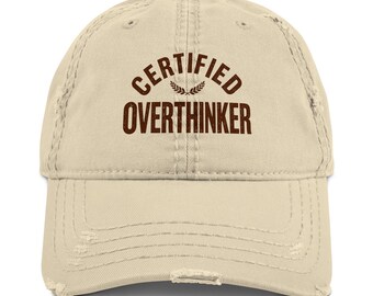 Könnte beinhalten: Eine beigefarbene Baseballkappe im Used-Look mit dem dunkelbraunen Schriftzug CERTIFIED OVERTHINKER, der über einem kleinen Lorbeerblatt-Symbol gewölbt ist. Die Kappe hat einen gebogenen Schirm mit ausgefransten Kanten und sechs Paneele.