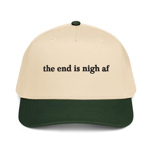 May include: A cream-colored baseball cap with a dark green brim and a dark green button on top. The front of the cap features the black embroidered text "the end is nigh af" in a serif font. The cap has a structured crown and stitched eyelets.