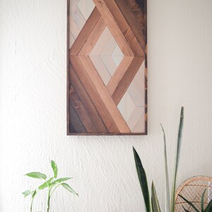 Puede incluir: Una pieza de arte de pared de madera geom&eacute;trica con un patr&oacute;n de diamante en tonos de marr&oacute;n, beige y blanco. El arte est&aacute; enmarcado en un marco de madera marr&oacute;n oscuro.