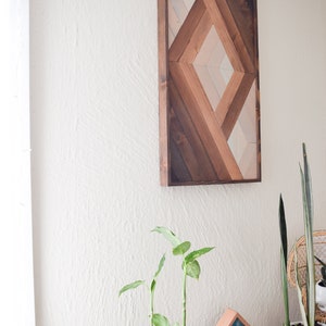 Puede incluir: Una pieza de arte de pared de madera geom&eacute;trica con un patr&oacute;n de diamante en tonos de marr&oacute;n, beige y blanco. El arte est&aacute; enmarcado en madera marr&oacute;n oscura y est&aacute; colgado en una pared blanca.