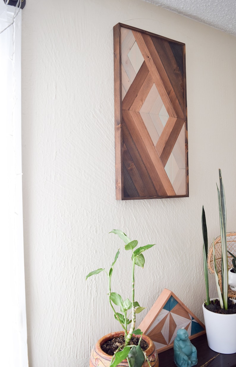 Puede incluir: Una pieza de arte de pared de madera geom&eacute;trica con un patr&oacute;n de diamante en tonos de marr&oacute;n, beige y blanco. El arte est&aacute; enmarcado en madera marr&oacute;n oscura y est&aacute; colgado en una pared blanca.