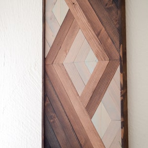Puede incluir: Una pieza de arte de pared geom&eacute;trica hecha de madera con un patr&oacute;n de diamante. La madera est&aacute; te&ntilde;ida en varios tonos de marr&oacute;n y blanco.
