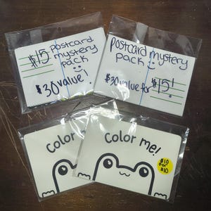 May include: Four rectangular postcard packs in clear plastic sleeves. Two top packs have handwritten text: $15 Postcard mystery pack $30 value! and Postcard mystery pack $30 value for $15!. Two bottom packs feature a frog illustration and text: color me!