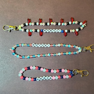 May include: Three beaded wristlet keychains in the image. The top keychain is black, white, and red with the name "HAKUJO" spelled out. The middle keychain is multi-colored with the name "KOYUKI". The bottom keychain is pink, blue, and white with the name "AKAZA".