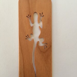 May include: A wooden bookmark with a cutout of a lizard in the shape of a silhouette. The bookmark is light brown in color.