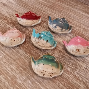 May include: Six ceramic teapot-shaped trinket dishes in various colors: red, blue, pink, and teal. Each dish has a unique glaze pattern, with a white base and colorful accents. The dishes are arranged on a wooden surface.