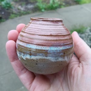 May include: A small, handmade ceramic vase with a rounded shape. The vase features horizontal bands of color, including shades of brown, pink, and blue. The vase is held in a hand, showcasing its size and texture. The pottery has a rustic, earthy aesthetic.