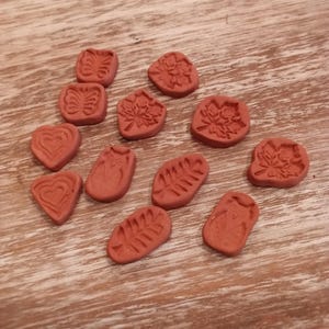 May include: A collection of small, reddish-brown clay stamps with various nature-inspired designs. The stamps feature shapes like butterflies, leaves, hearts, and owls. They are scattered on a light brown wooden surface.