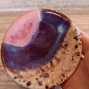 May include: A round ceramic coaster with a glossy finish. The coaster features a blend of colors, including pink, purple, and beige, with dark brown speckles along the edge. The coaster is approximately 4 inches in diameter.