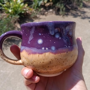May include: A ceramic mug featuring a two-tone glaze design. The upper portion is a deep purple with lighter blue and white speckled accents, while the lower portion is a speckled tan color. The mug has a curved handle and a rounded body shape.