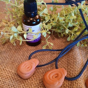 May include: Two terracotta-colored ceramic pendants shaped like cinnamon rolls, hanging from a dark blue cord. A small bottle of essential oil with a black cap is in the background.