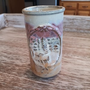 May include: A handmade ceramic tumbler with a landscape design. The tumbler features a light beige base with a reddish-purple band near the top and a gray rim. The landscape design includes trees and a house, all within an oval frame.