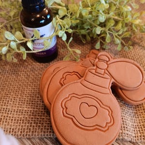May include: Three terracotta-colored ceramic bottle-shaped ornaments with a heart design. The ornaments are on a burlap surface next to a small bottle of lavender essential oil.