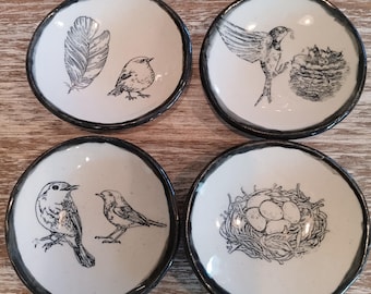 May include: Set of four small, round ceramic dishes with a white interior and a dark gray rim. Each dish features a different black and white illustration of birds, nests, or feathers. The dishes are arranged on a light wood surface.