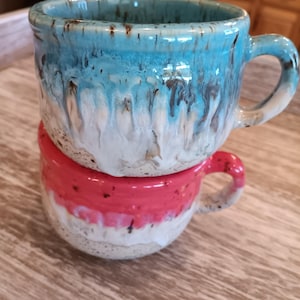 May include: Two ceramic mugs stacked on top of each other. The top mug is light blue with white and brown streaks. The bottom mug is white with a pink stripe and brown speckles.