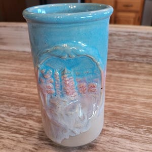 May include: A handmade ceramic tumbler with a light blue glaze. The tumbler features a raised relief design of a cityscape in shades of pink and white, framed by a decorative border. The bottom portion is a natural, unglazed color.