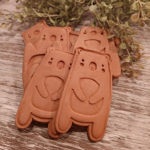 May include: Five terracotta bear-shaped ornaments with a simple, smiling face design. The ornaments are arranged in a pile on a wooden surface.