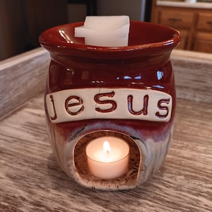 May include: A red and white ceramic wax warmer with the word "Jesus" on the front. A white wax melt is in the top opening and a small white tea light is in the bottom opening.