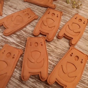 May include: Seven terracotta bear-shaped ornaments with a simple design. The bears are standing upright with their arms raised and have a small, round head with two small dots for eyes.