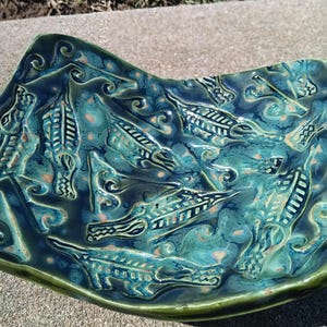 May include: A rectangular ceramic dish with a glossy teal and dark blue glaze. The surface features a repeating embossed pattern of stylized alligator figures and swirling wave-like textures throughout the entire interior of the piece.