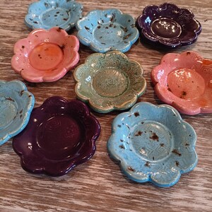 May include: A collection of small, flower-shaped ceramic dishes in various colors, including blue, pink, purple, and green. Each dish has a central well and a scalloped edge, with a rustic, speckled finish. The dishes are arranged on a wooden surface.