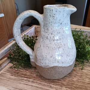 May include: A white ceramic pitcher with brown speckles and a brown base. The pitcher has a wide mouth and a handle.