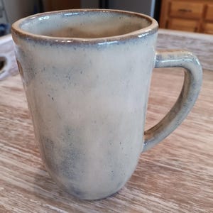 May include: A handmade ceramic mug with a rounded body and a curved handle. The mug has a speckled, off-white glaze with blue and brown accents. The interior and rim are a darker shade of brown.
