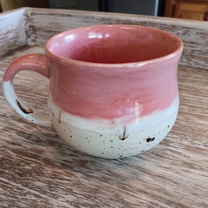 May include: A ceramic mug with a rounded shape and a curved handle. The upper portion of the mug is glazed in a soft pink, while the lower portion is a creamy off-white with dark brown speckles scattered across the surface. The interior is pink.