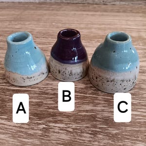 May include: Three ceramic inkwells, one with a purple glaze and two with a light blue glaze. Each inkwell has a white base with speckles.
