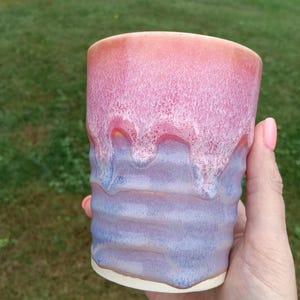 May include: A ceramic tumbler featuring a smooth, glossy glaze with a gradient transition from a soft coral pink at the top to a muted lavender blue at the base. The surface has a subtle rippled texture with white drip-like glaze accents.