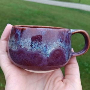 May include: A ceramic mug with a rounded shape and a handle, featuring a deep purple and maroon glaze with a textured, drippy blue and white crystalline pattern around the upper rim and a smooth, solid dark purple base.