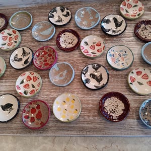 May include: A collection of 20 small ceramic dishes with various designs. The dishes feature images of chickens, strawberries, lemons, and other patterns. The dishes are all hand-painted and have a rustic, vintage look.