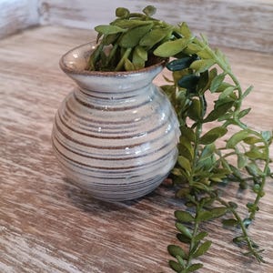 May include: A small, round ceramic vase with horizontal brown and white stripes. The vase is filled with green leafy faux plants. The vase has a narrow neck and a slightly flared rim. The vase is sitting on a wooden surface.