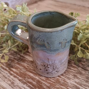 May include: A small, green ceramic pitcher with a brown and white glaze. The pitcher has a handle and a spout.