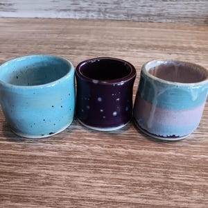 May include: Three ceramic mugs with different glaze patterns. The first mug is light blue with speckles, the second is dark purple with white speckles, and the third is light blue and pink with a white glaze.