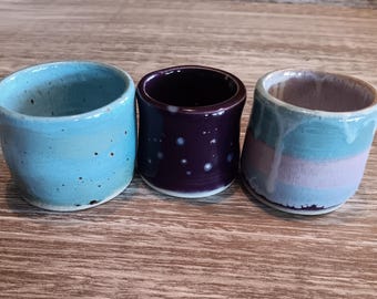 May include: Three ceramic mugs with different glaze patterns. The first mug is light blue with speckles, the second is dark purple with white speckles, and the third is light blue and pink with a white glaze.