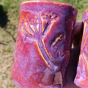 May include: A cylindrical ceramic mug featuring a vibrant, mottled glaze in shades of deep magenta, purple, and orange. The surface is decorated with a raised, embossed botanical pattern of a flowering plant with delicate, branching stems.