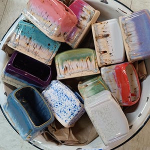 May include: A collection of small, colorful ceramic planters with various patterns and glazes. The planters are in a white enamel bowl.