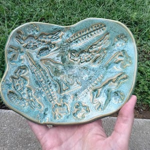 May include: An irregular, oval-shaped ceramic dish with a glossy, mottled turquoise and teal glaze. The surface features recessed, intricate relief patterns of various botanical and abstract shapes, with gold-toned accents highlighting the textured details.