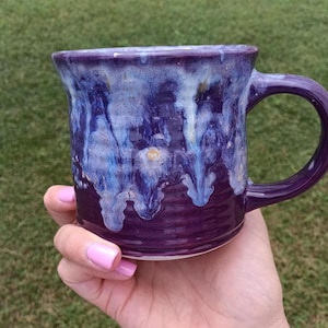 May include: A ceramic mug with a deep purple base and a glossy glaze featuring white and light blue drips cascading from the rim. The exterior surface has a subtle horizontal ribbed texture, and the mug includes a curved handle on the right side.