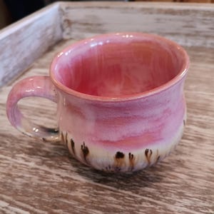 May include: A handmade ceramic mug with a pink and white glaze. The mug has a rounded shape with a handle and a decorative pattern of brown brushstrokes near the base. The mug is designed for hot beverages.