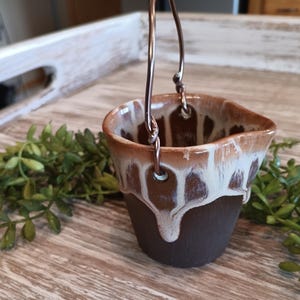 May include: A small, handmade ceramic bucket with a copper wire handle. The bucket is dark brown with a white and brown glaze dripping down the sides. The bucket is sitting on a wooden surface.