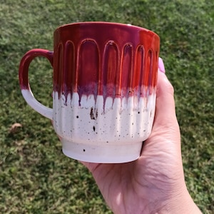 May include: A ceramic mug featuring a vertical ribbed texture with a glossy deep red glaze on the upper portion that drips down into a speckled off-white base. The mug includes a curved handle on the side and a smooth, cylindrical body shape.