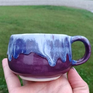 May include: A ceramic mug with a rounded body and a side handle, featuring a deep purple base glaze that transitions into a white drip pattern near the rim. The interior and the bottom of the mug are finished in a smooth, solid white glaze.
