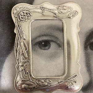 May include: A small, ornate silver-toned picture frame with a rectangular opening. The frame is decorated with floral and leaf motifs. The image within the frame is a black and white close-up of an eye.