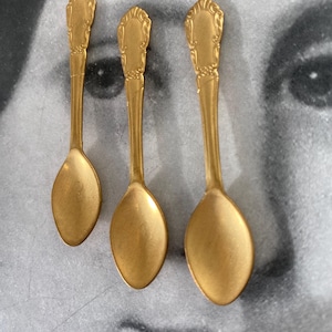 May include: Three small, gold-colored spoons with ornate, decorative handles. The spoons have oval-shaped bowls and are arranged on a gray background. The spoons are likely for serving or display purposes.