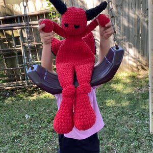 May include: A red crocheted devil plush toy with black horns and eyes. The toy is being held by a child on a swing set.